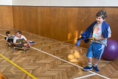 Žáci v tělocvičně s hrají s pojízdnou deskou a vozí se na ní.